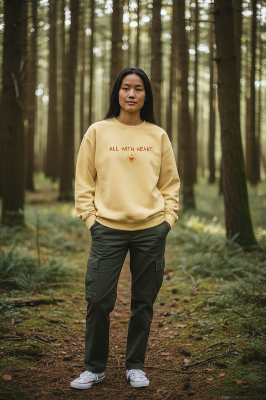 A woman wearing in the forest wearing a yellow Crewneck that says “All With Heart” in embroidery.