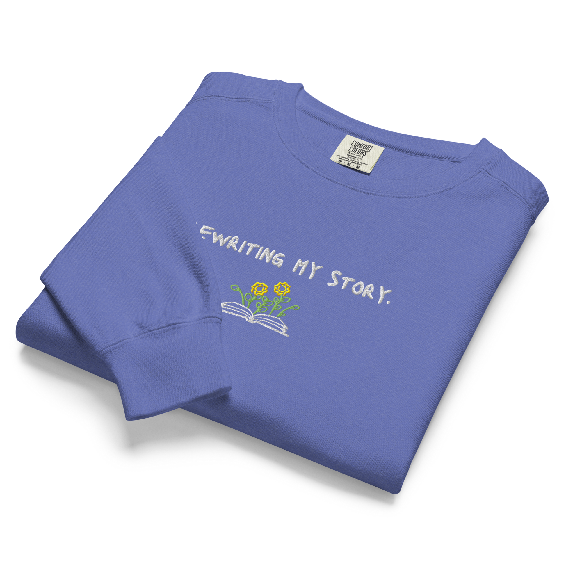 A folded blue Crewneck that says “Rewriting My Story” with an open book and flowers design.