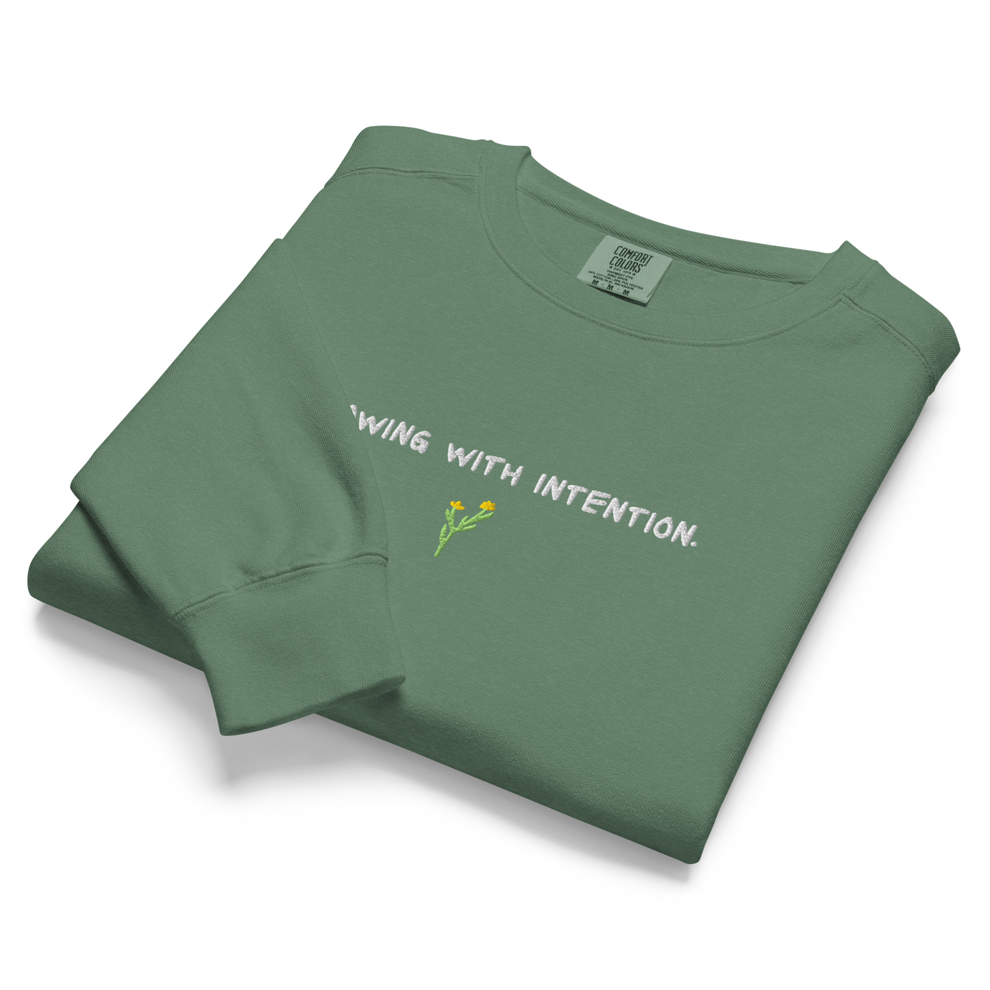 A folded green eco-conscious crewneck with “Growing With Intention” text and yellow flower design.