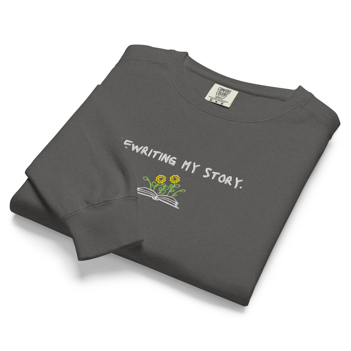 A folded pepper-colored Crewneck that says “Rewriting My Story” with an open book and flowers design.